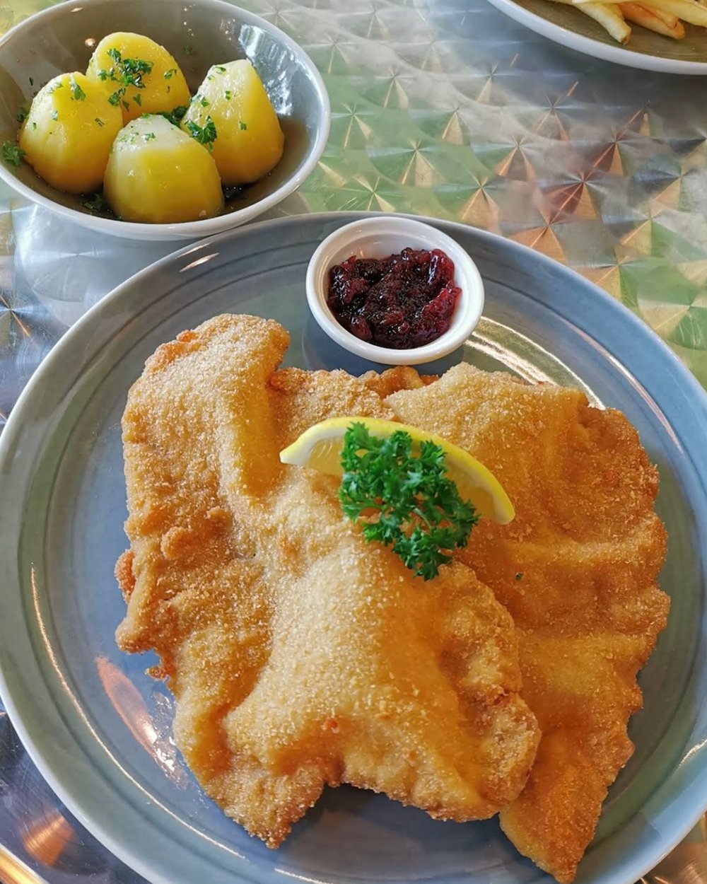 Wiener Schnitzel on a stone-blue plate with lemon, parsley, a bowl of parsley potatoes and a pot of lingonberries