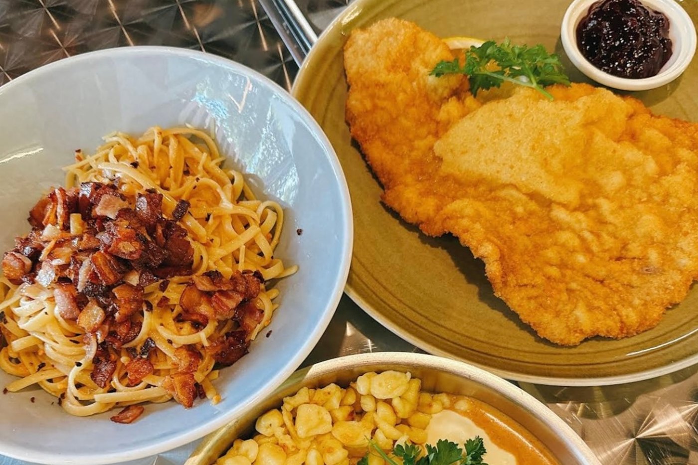 Three plates on a steel table: bacon linguine, a Wiener Schnitzel with lingonberries, and a schnitzel with Spaetzle